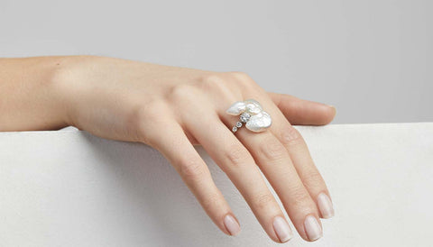 A woman's hand rests on a white surface, she wears a diamond ring which has a pearl oozing around it